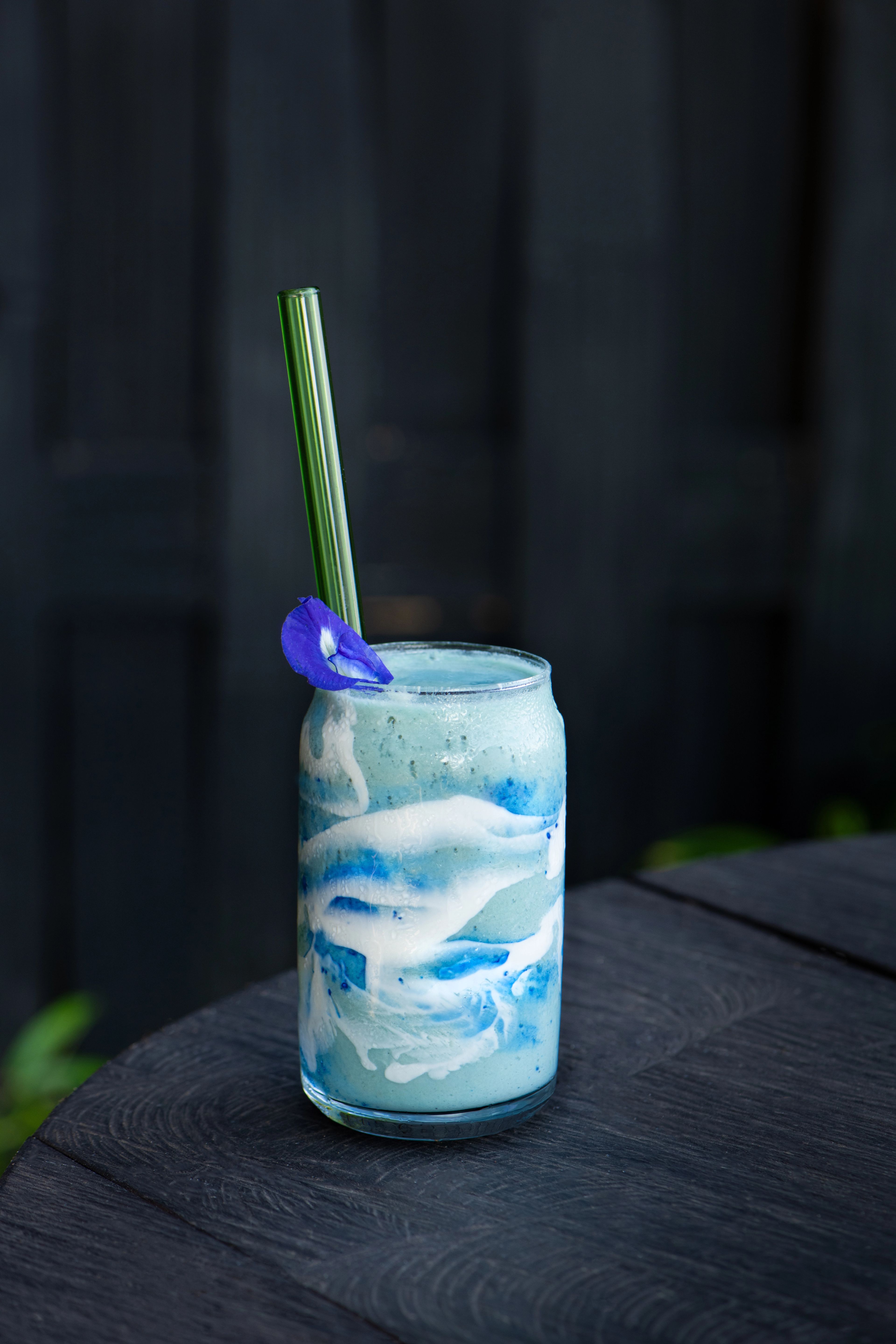 A blue and white layered butterfly pea drink in a tumbler with a glass straw and a butterfly pea flower on the rim, on a dark timber table.