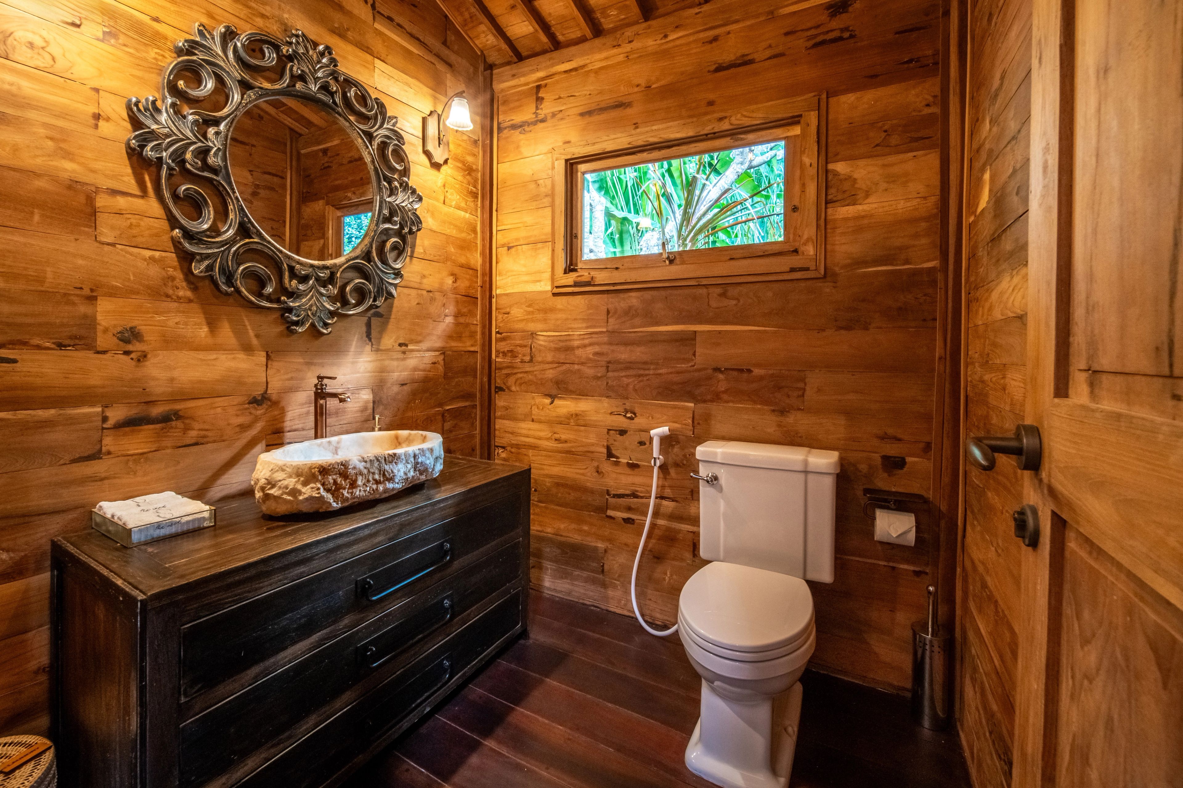 A stone basin on a dark chest below a carved round mirror, in a timber-lined bathroom.