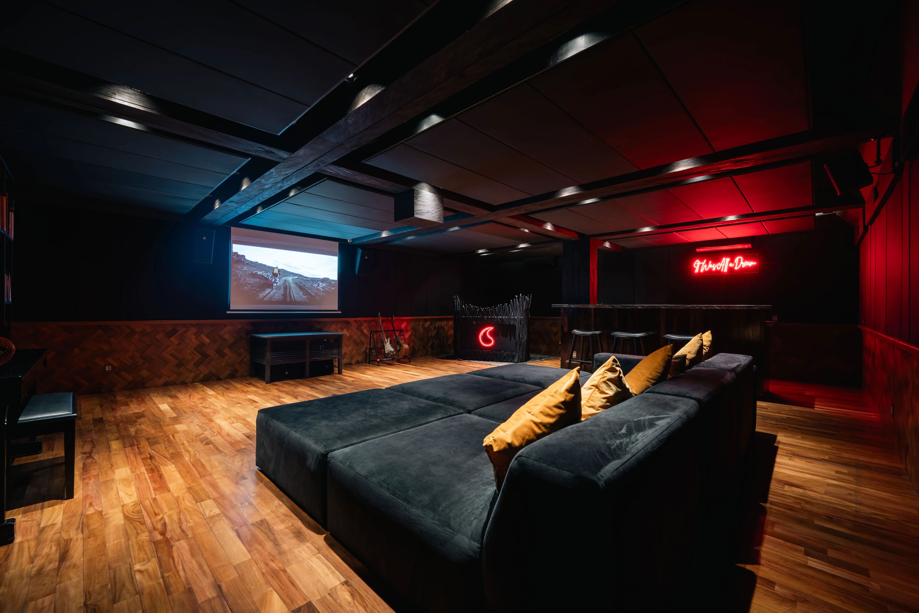 Deep sofas facing the screen in the basement, the bar's red neon glowing at the far end.
