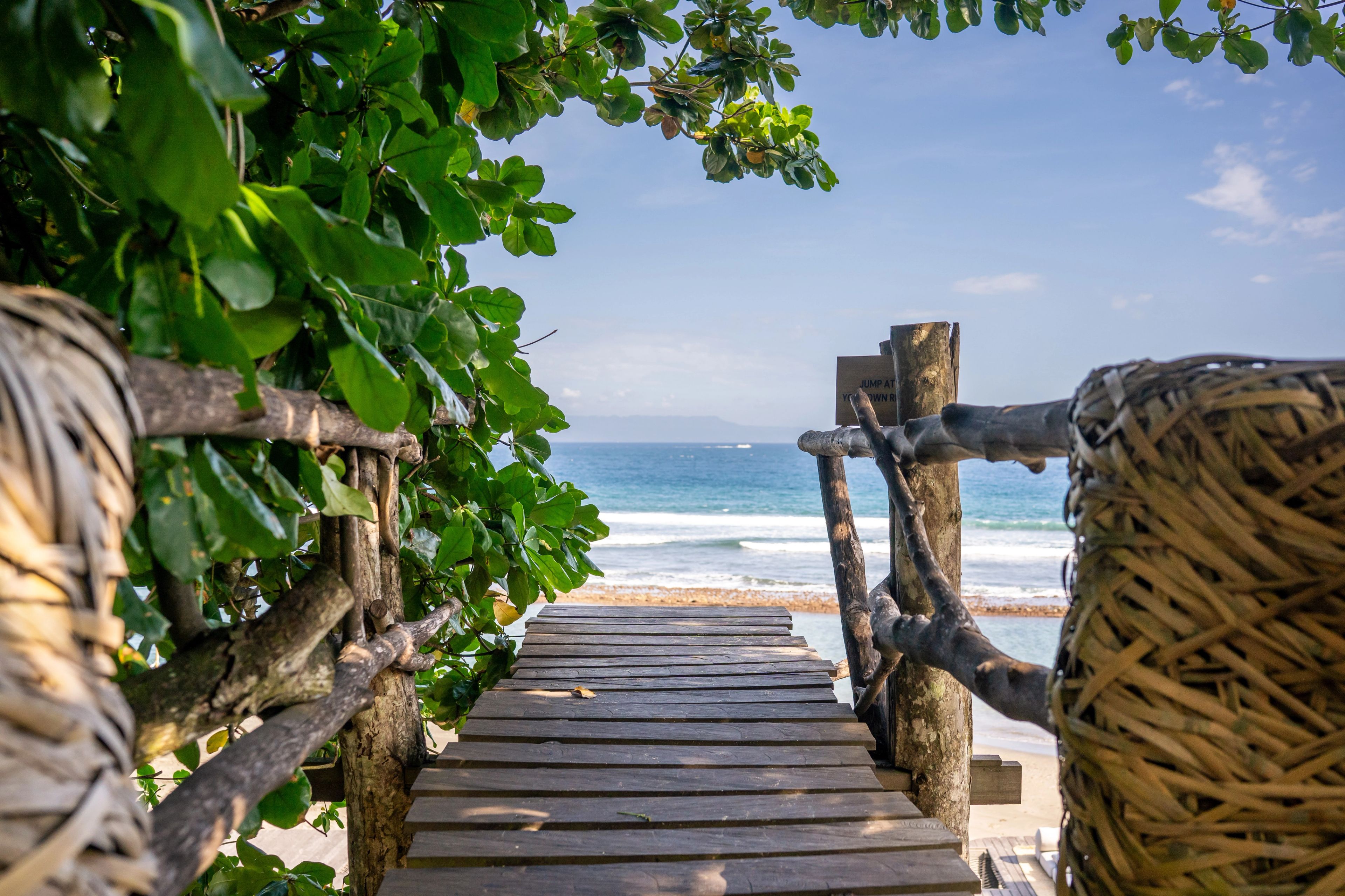 A timber boardwalk framed by leaves and a woven pod, running down towards the shore.