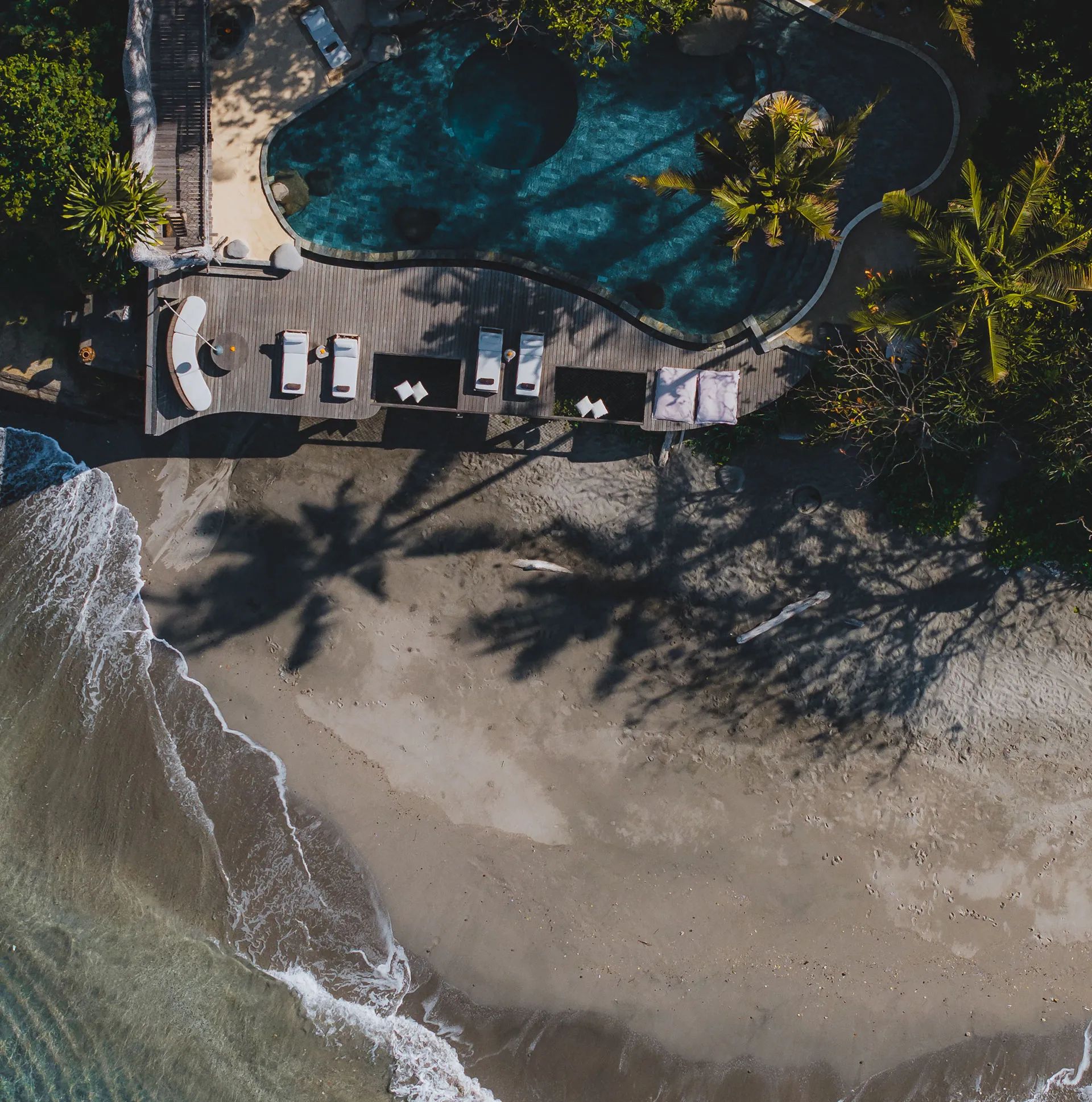 An aerial view straight down onto the timber deck and its daybeds, the surf breaking on the sand beside it.