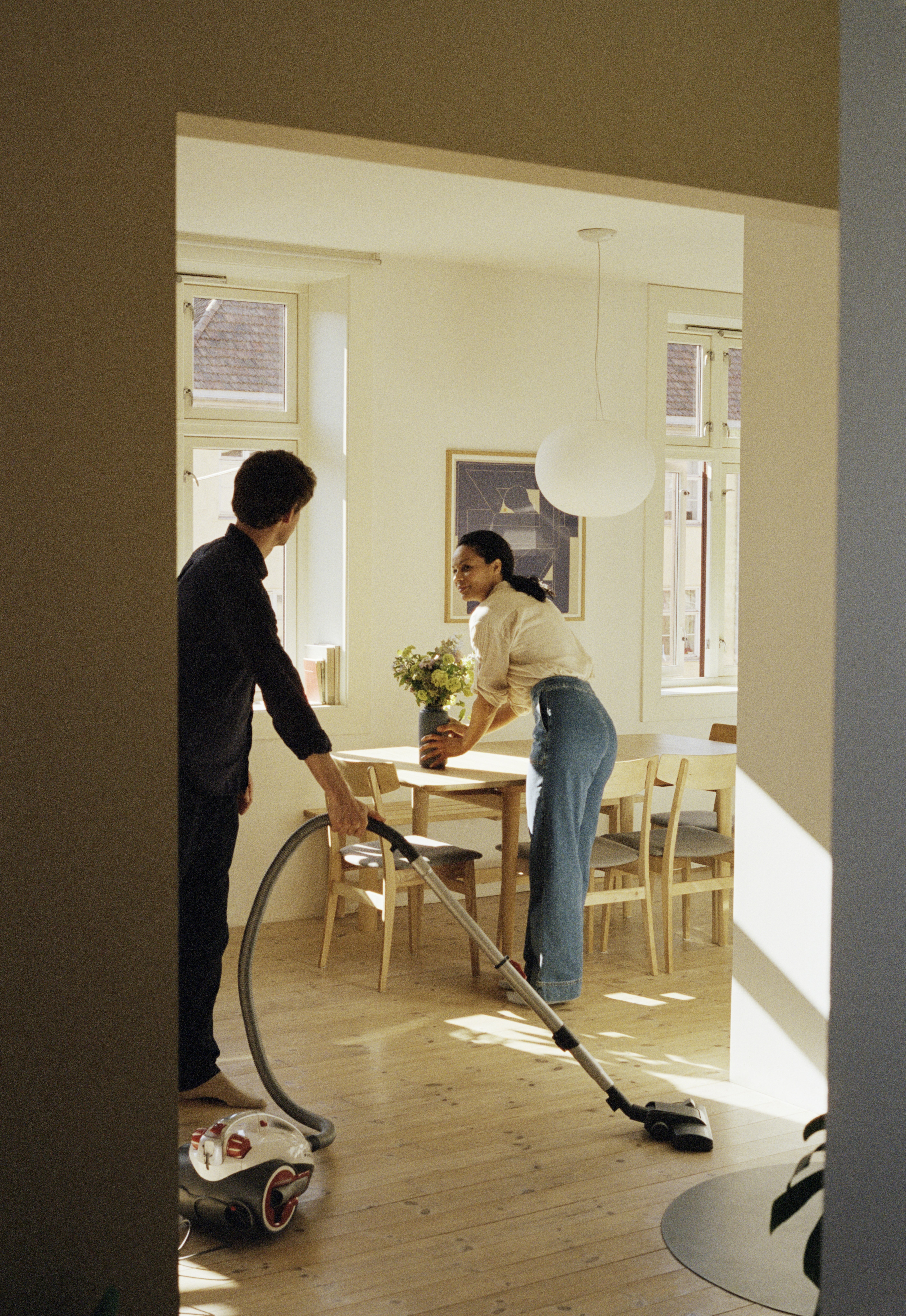 A person vacuums a wooden floor while another arranges flowers in a vase on a dining table in a well-lit room.