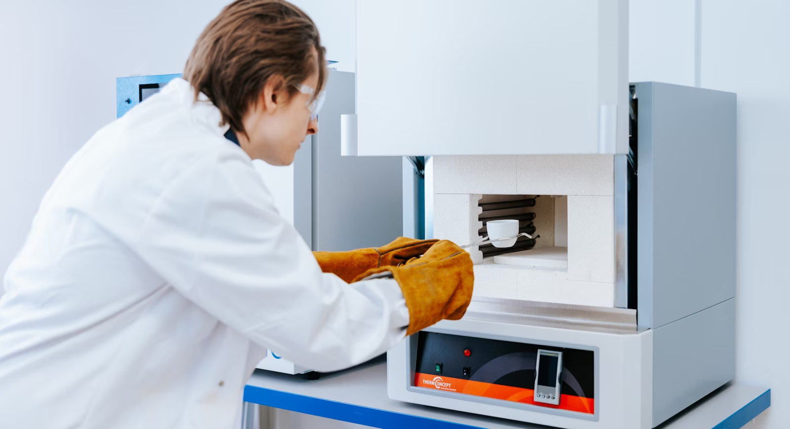 A Photoncycle lab scientist in protective gear places a ceramic sample into a high-temperature furnace, revealing its insulated interior.