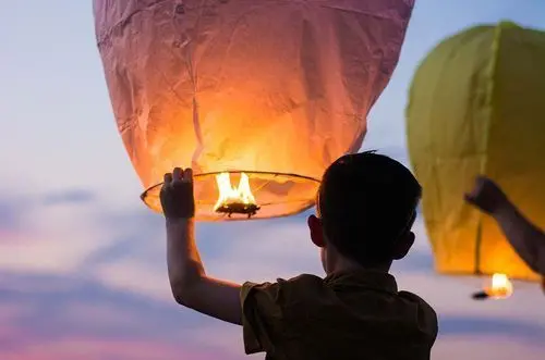 A child, seen from behind, holds up a pink paper lantern as the flame inside ignites and fills it with a warm, orange glow, preparing it for release into the dusk sky, with a second, yellow lantern visible on the right.