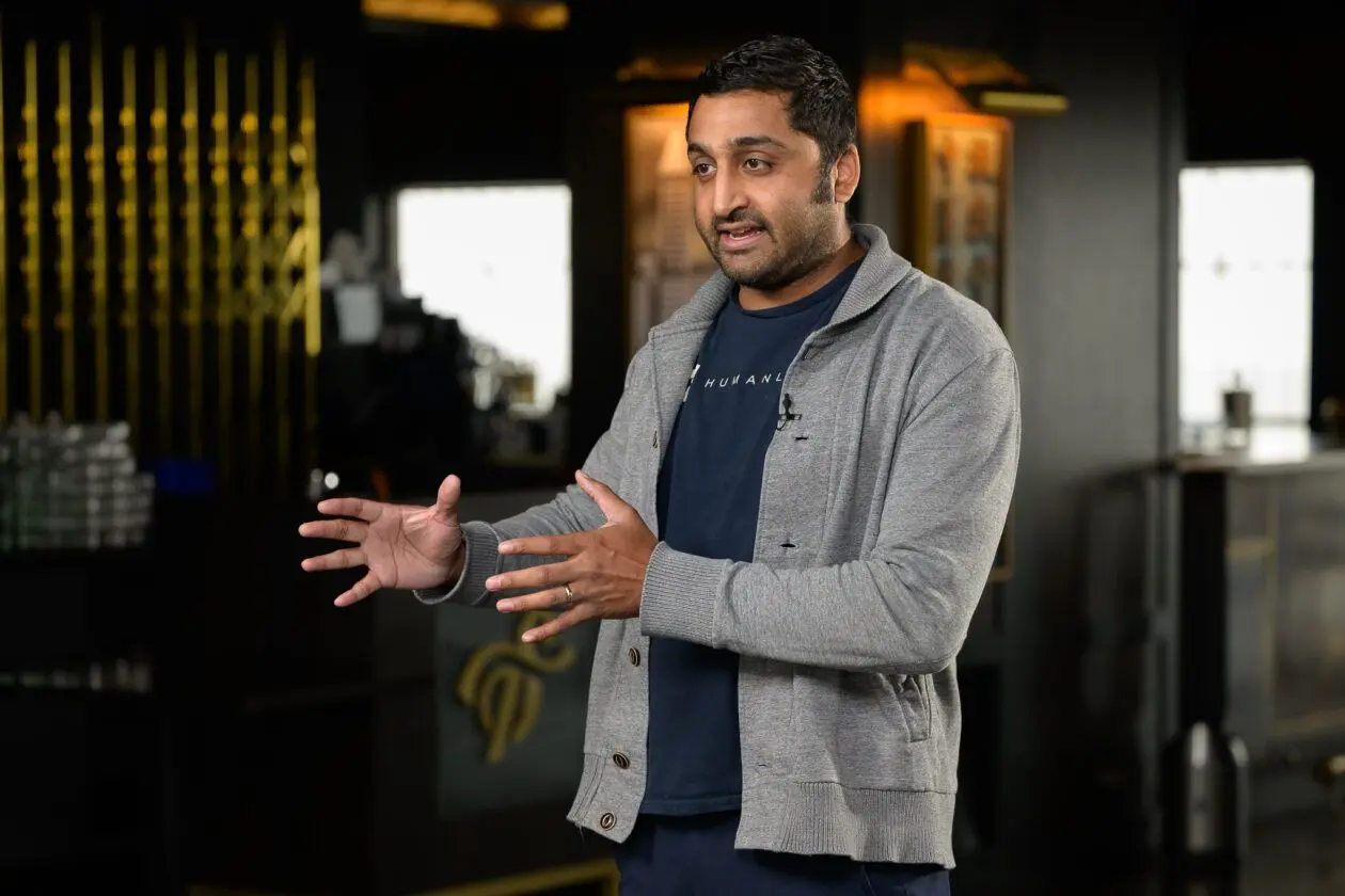 Person speaking and gesturing with hands while wearing a grey sweater and a Humanly t-shirt, in a modern indoor setting.