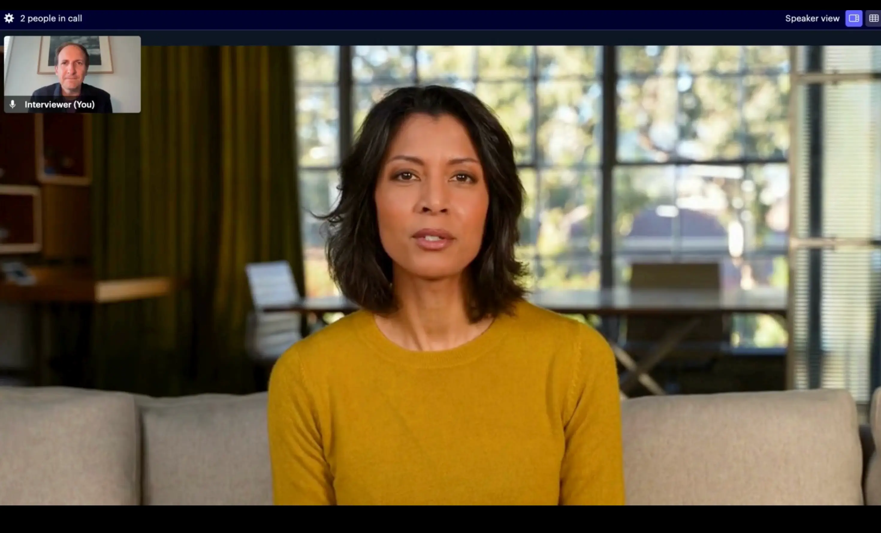 A screenshot of a two-person video conference call interface. The main view features a woman with dark hair wearing a mustard yellow sweater, speaking while seated on a light-colored couch in a well-lit room with large windows in the background. A small inset window in the top left shows the other participant, labeled "Interviewer (You)".