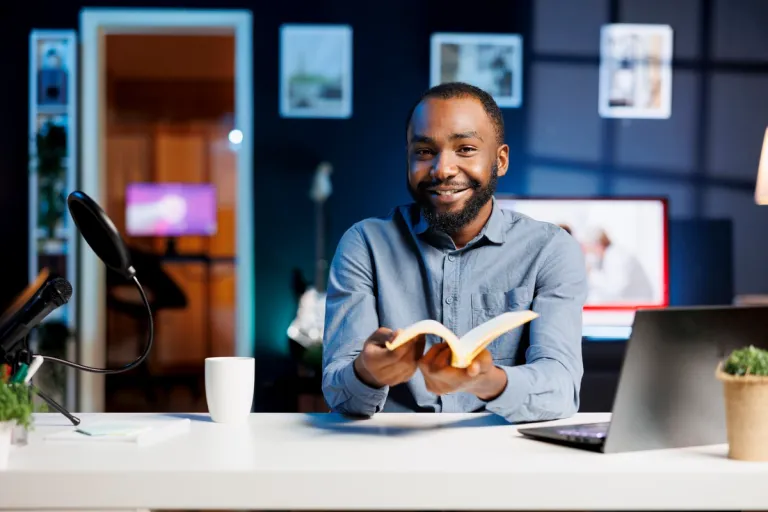 An author smiling at the camera while holding an open book at a desk with a microphone and laptop, ready to record a video