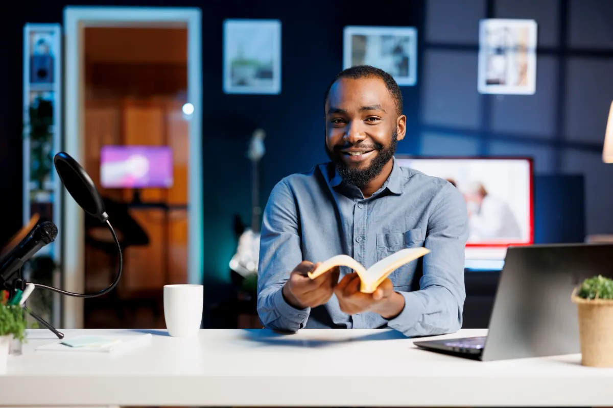 A smiling author sitting at a desk holding an open paperback book, with a microphone and laptop beside him