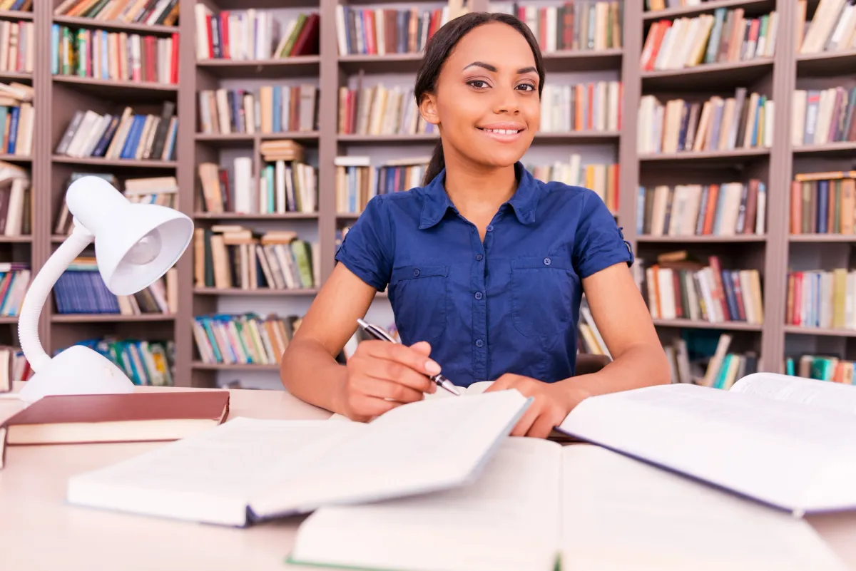 A smiling young woman writing notes at a desk covered with open books in front of tall bookshelves