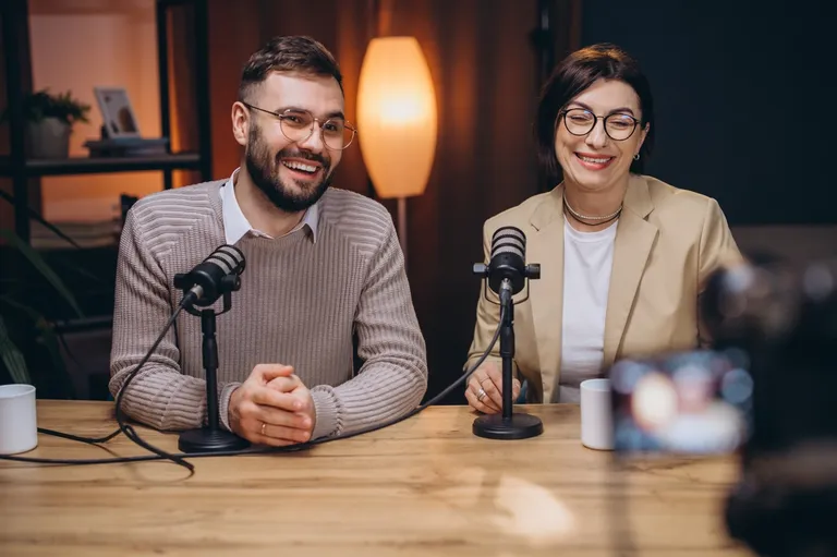 Two people smiling while speaking into studio microphones at a wooden table, ready to record audio