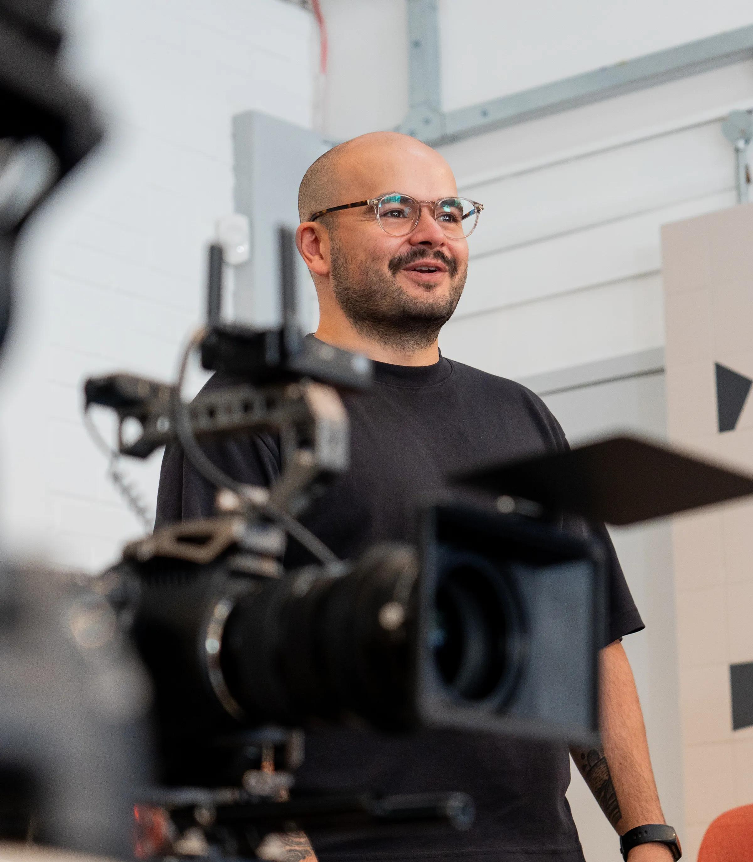 A bald man with glasses and a beard smiles while being filmed by a camera.