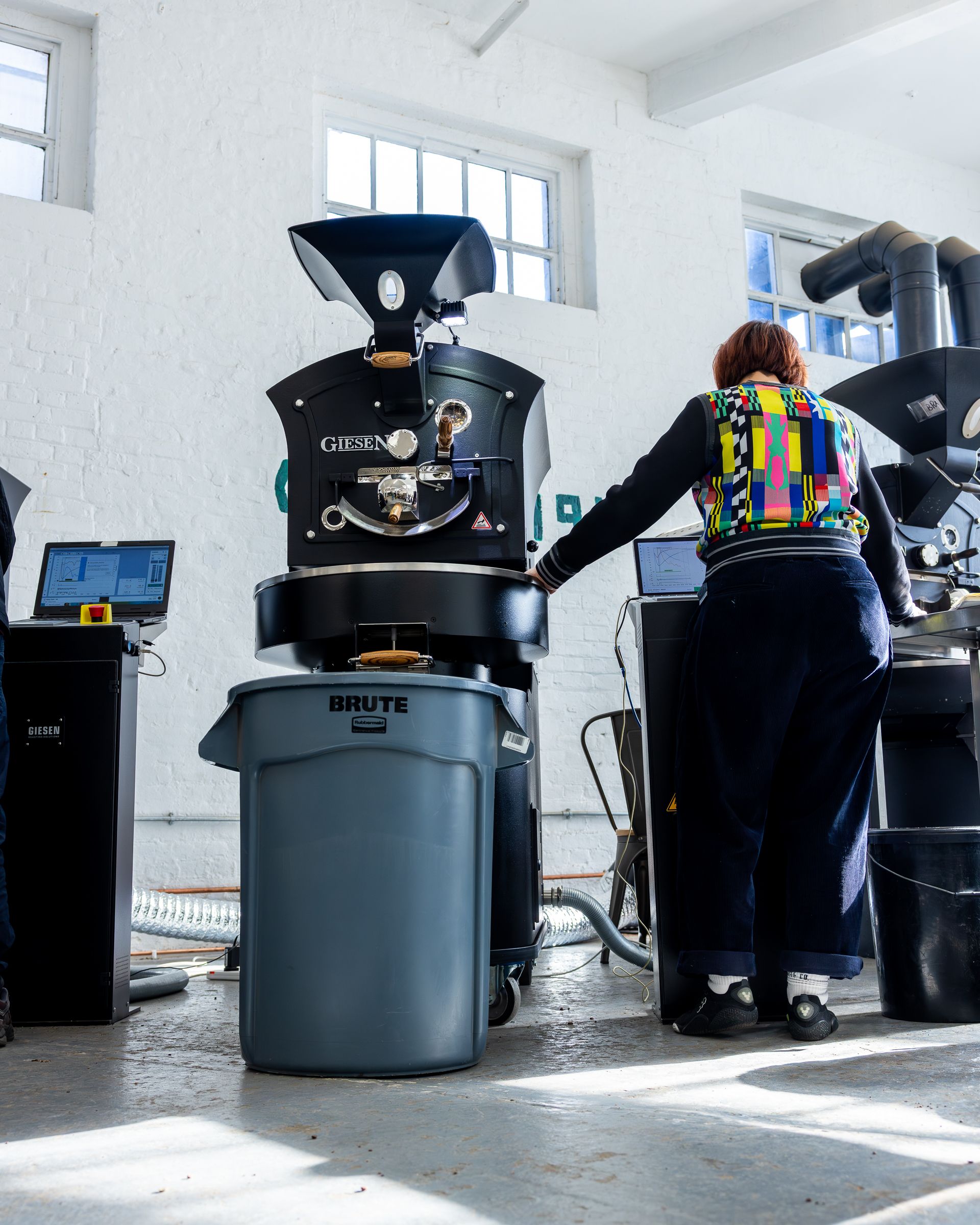 A person in a colorful jacket operates a Giesen coffee roaster in a bright, white-walled facility.