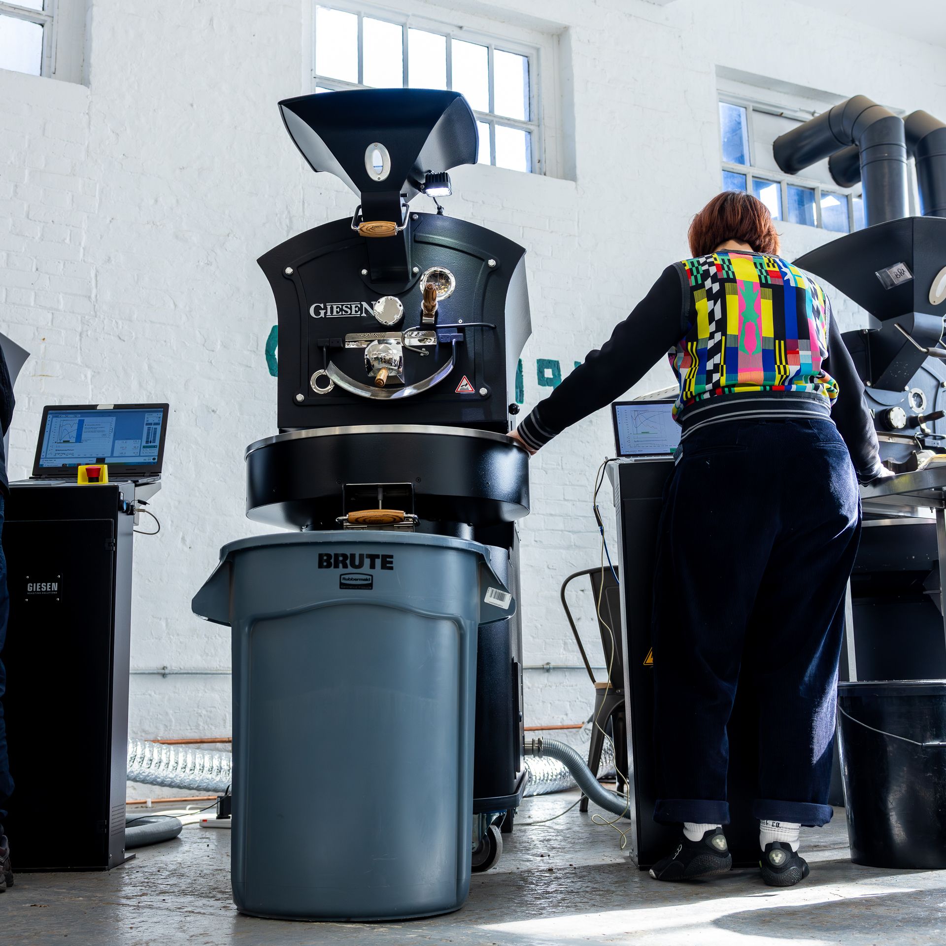 A person in a colorful jacket operates a Giesen coffee roaster in a bright, white-walled facility.