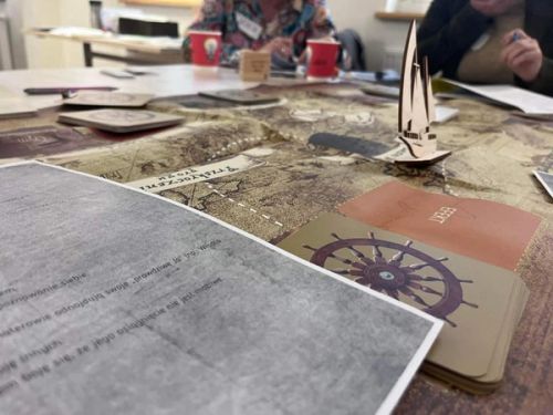 Bliski kadr na stół szkoleniowy, na którym rozłożona jest plansza przypominająca starą mapę. Na pierwszym planie widoczne są karty (jedna z grafiką koła sterowego, druga brązowa z napisem „EFEKT”) oraz wydrukowane materiały szkoleniowe. Na mapie stoi drewniana miniaturka żaglówki, a w rozmazanym tle widać siedzących uczestników warsztatów.