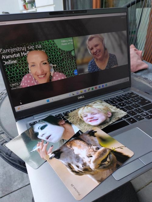 Laptop marki Dell z włączonym spotkaniem online na platformie Zoom, na którym widać dwie uśmiechnięte kobiety. Na klawiaturze laptopa leżą trzy drukowane karty Points of You (Punctum): uśmiechnięte dziecko w piasku, osoba z białą makijażem scenicznym paląca papierosa oraz zbliżenie na pysk wielbłąda