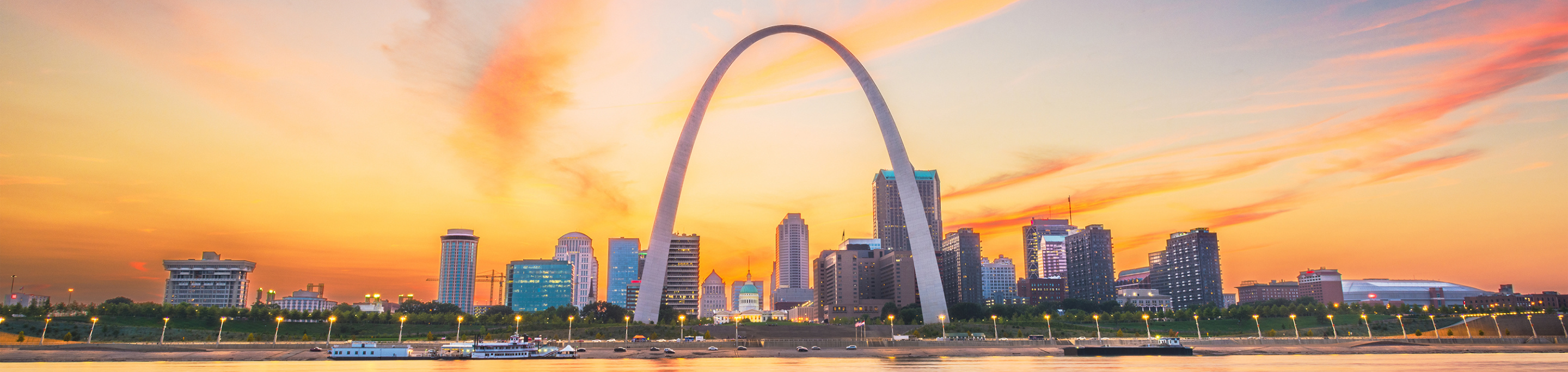 The Gateway Arch in closeup with a blurred background of the St. Louis skyline illuminated by a colorful sunset in Missouri.