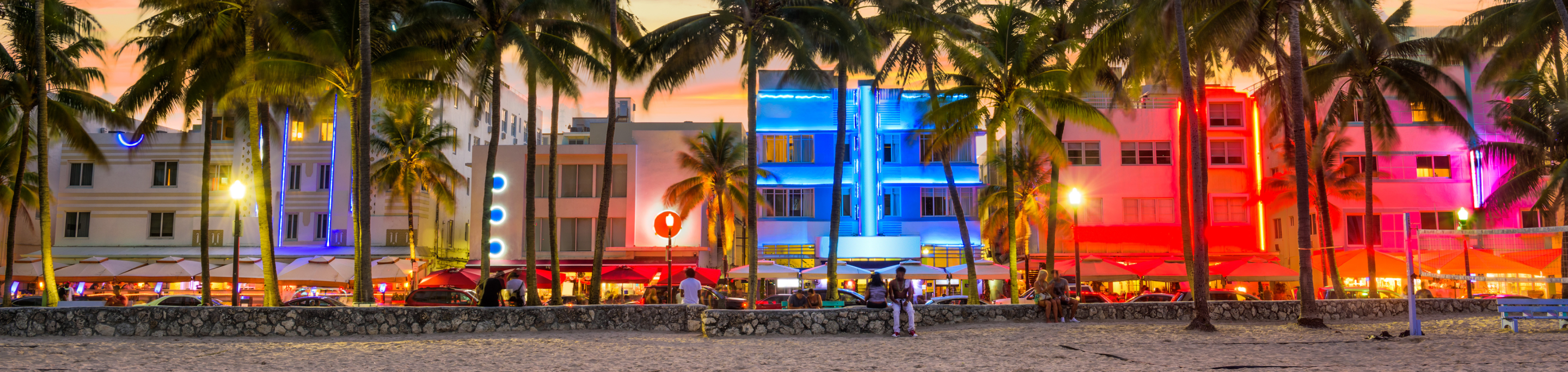 Vibrant neon-lit buildings in closeup with a blurred background of palm trees and a sandy beach at sunset in Miami's South Beach, Florida.