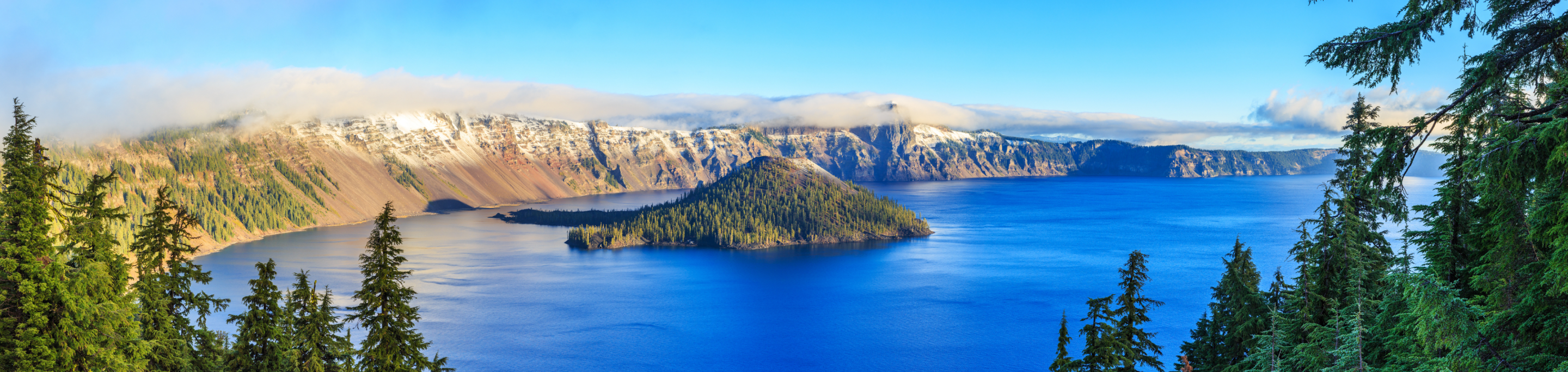 A panoramic view of Crater Lake in closeup with a blurred background of rugged cliffs, snow-dusted peaks, and evergreen trees under a bright blue sky in Oregon.