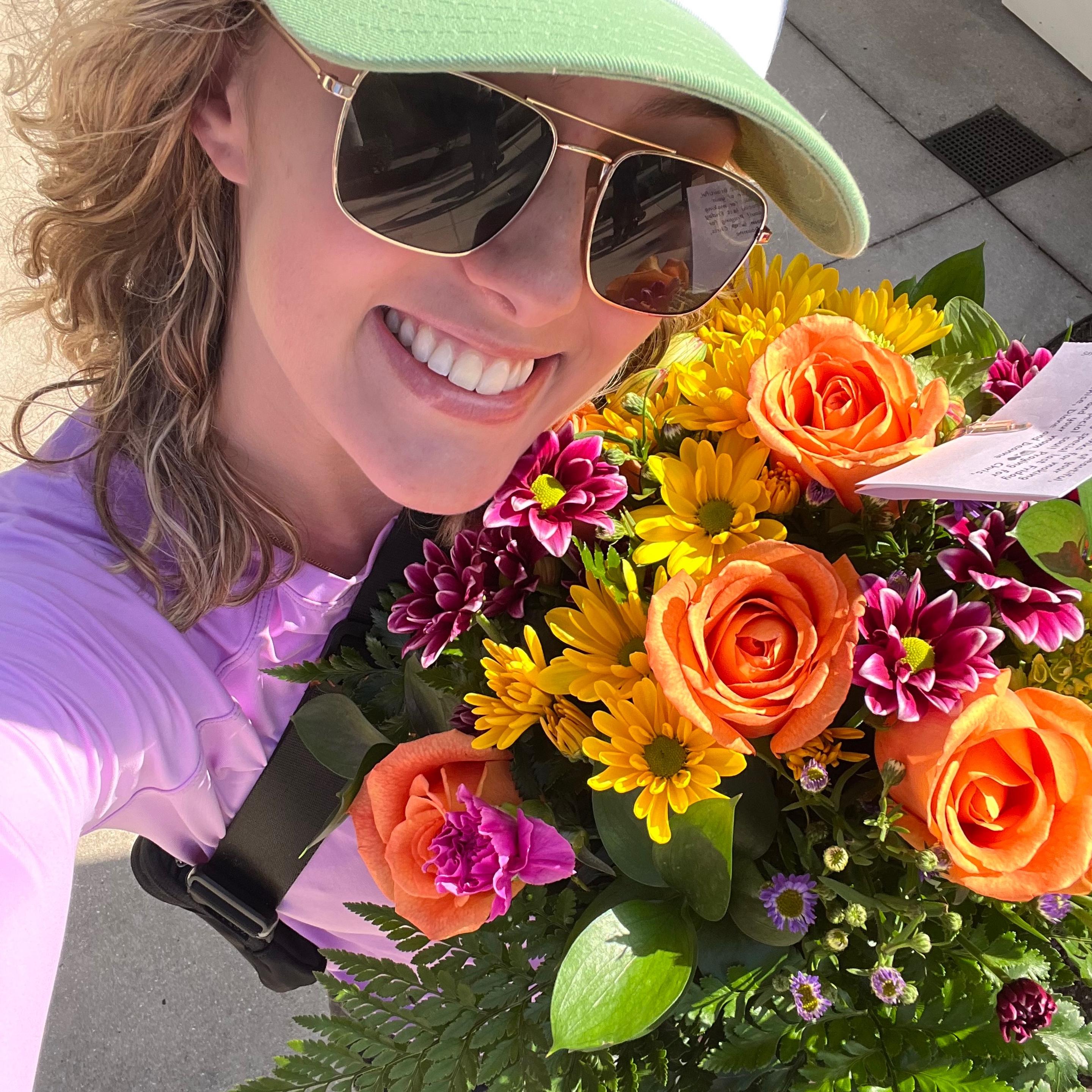 Woman in hat and sunglasses smiling, holding a bright orange flower arrangement