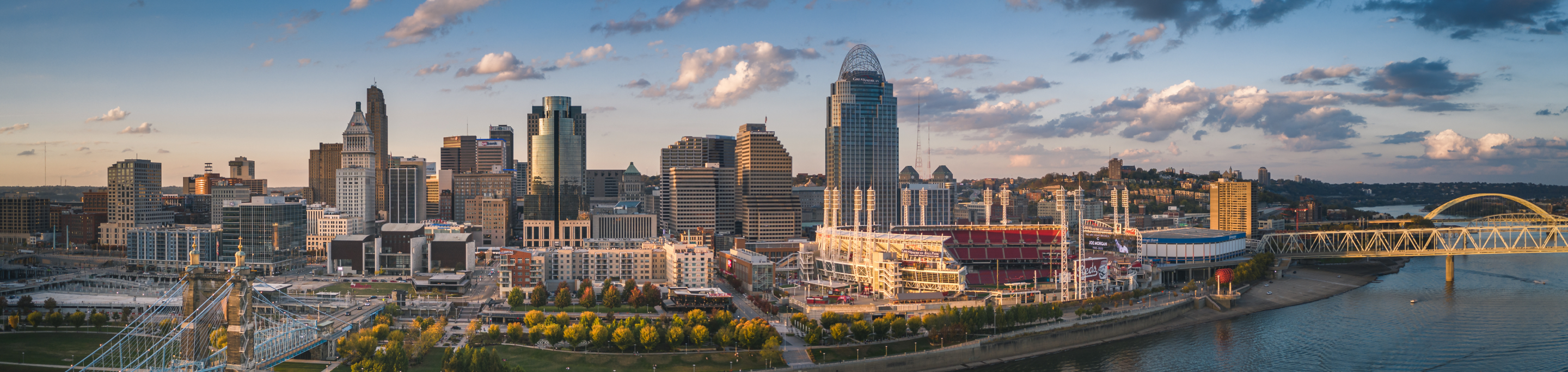 A panoramic view of Ohio's bustling city skyline, with towering buildings reflecting the soft light of the early evening, framed by the river and bridges, all under a sky dotted with scattered clouds.