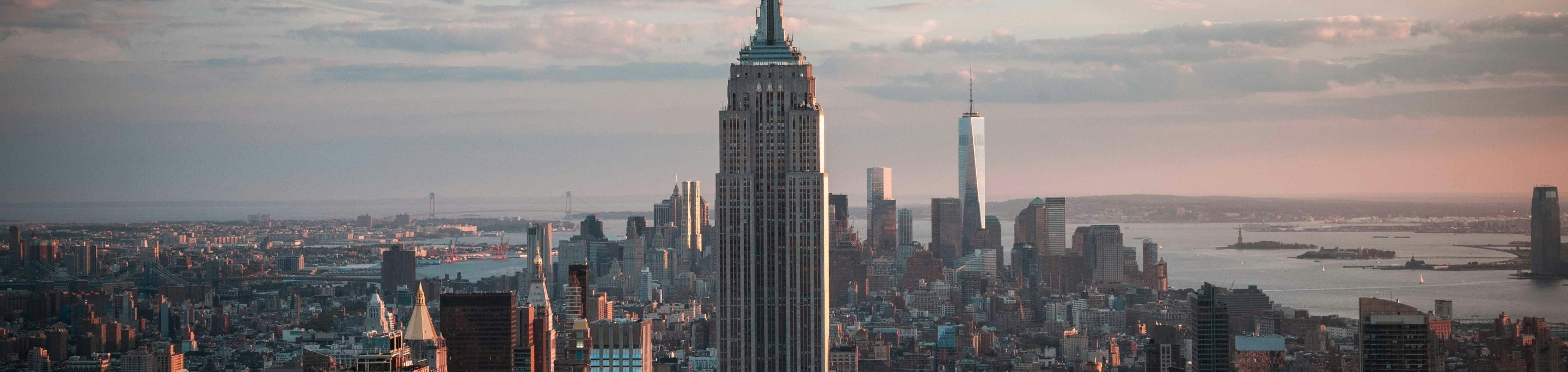 The Empire State Building in closeup with a blurred background of the New York City skyline and the Statue of Liberty in the distance under a soft evening light in New York.