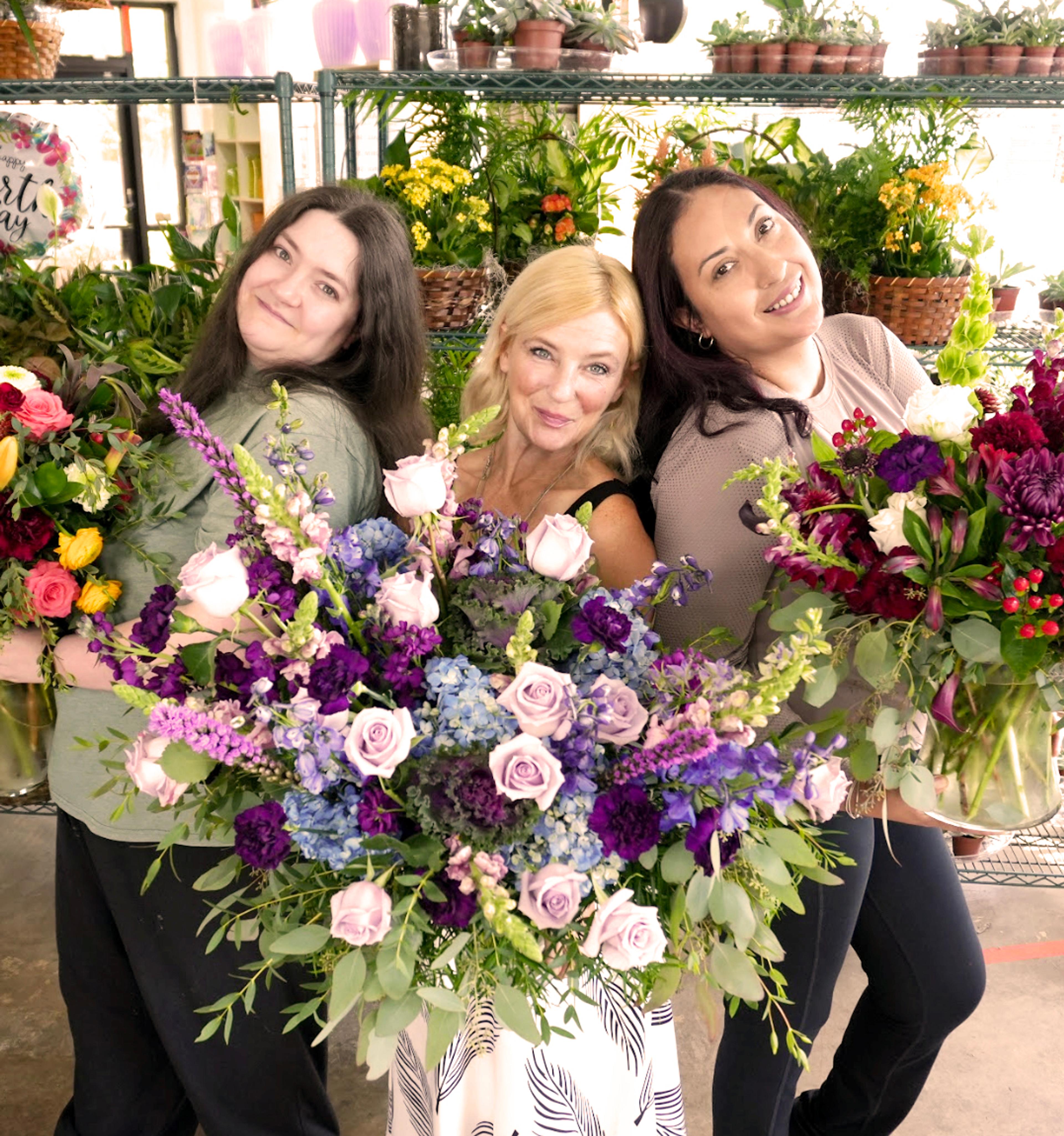 Three smiling women hold large, colorful floral bouquets in a flower shop.