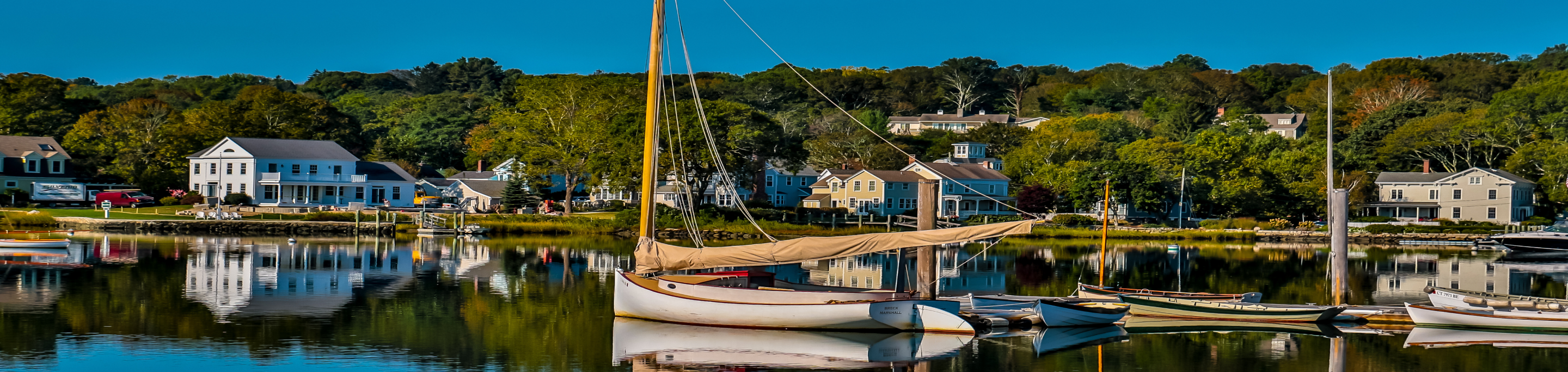 A sailboat docked in closeup with a blurred background of charming coastal homes and lush trees reflecting on the calm water in Mystic, Connecticut.