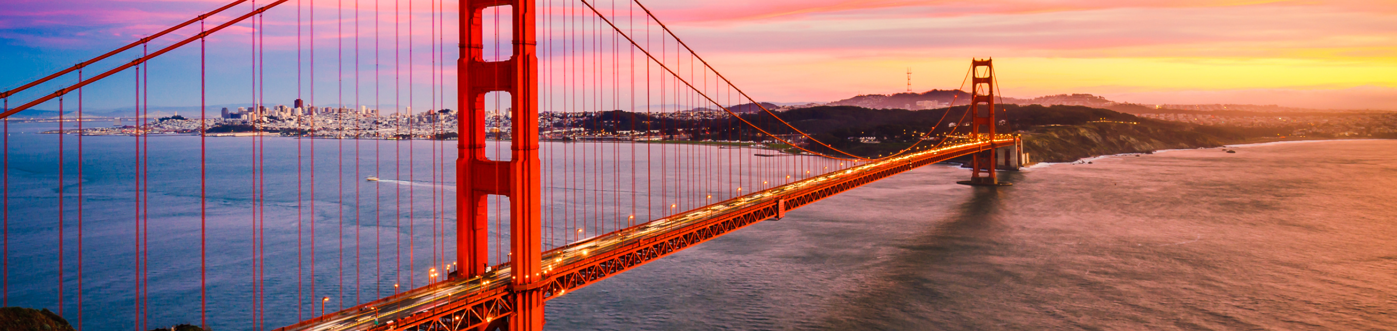 The Golden Gate Bridge in closeup with a blurred background of the San Francisco skyline and a vibrant sunset over the bay in California.