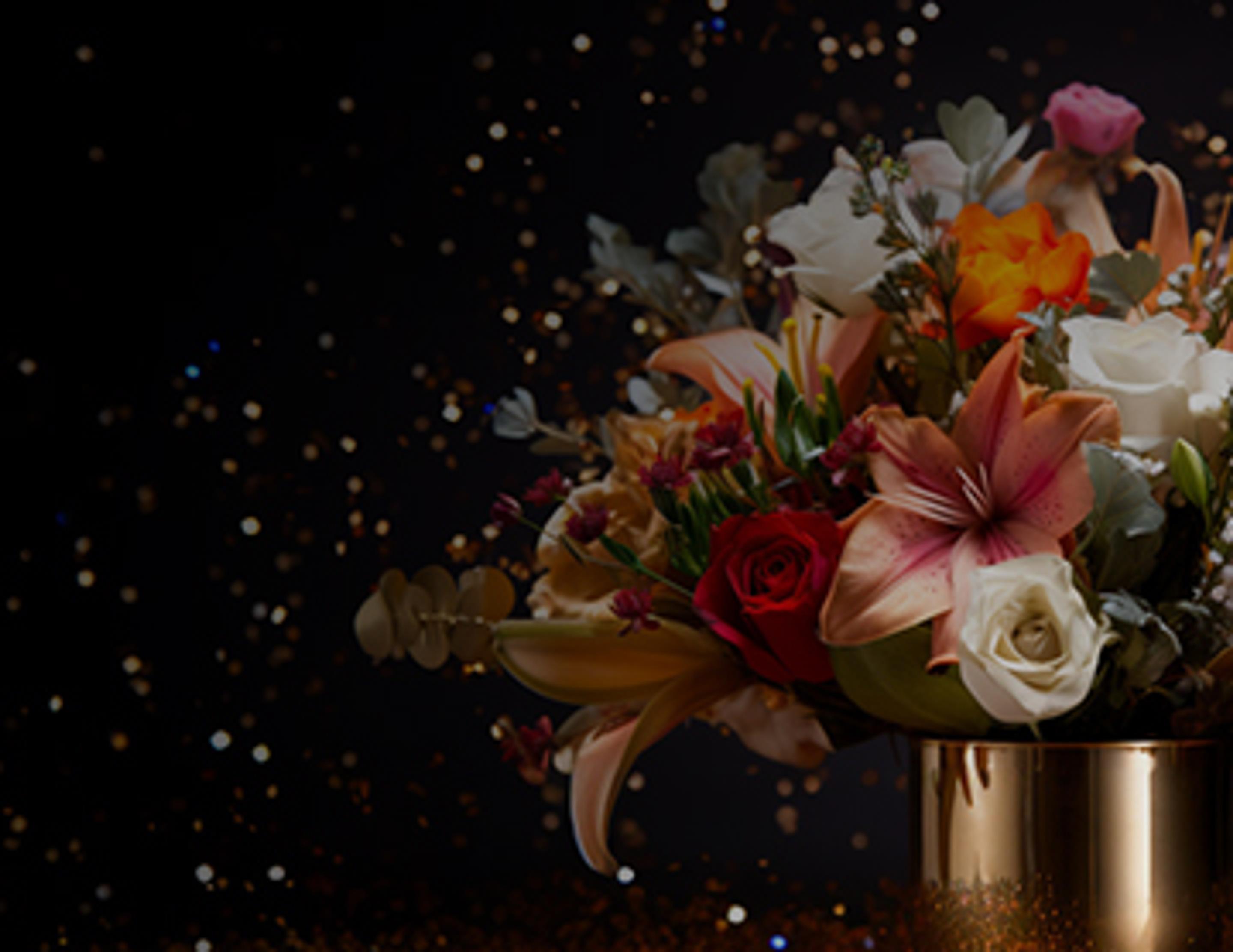 Luxurious floral arrangement in a golden vase featuring a rich mix of lilies, roses, and other assorted flowers, set against a dark backdrop with glittering accents.