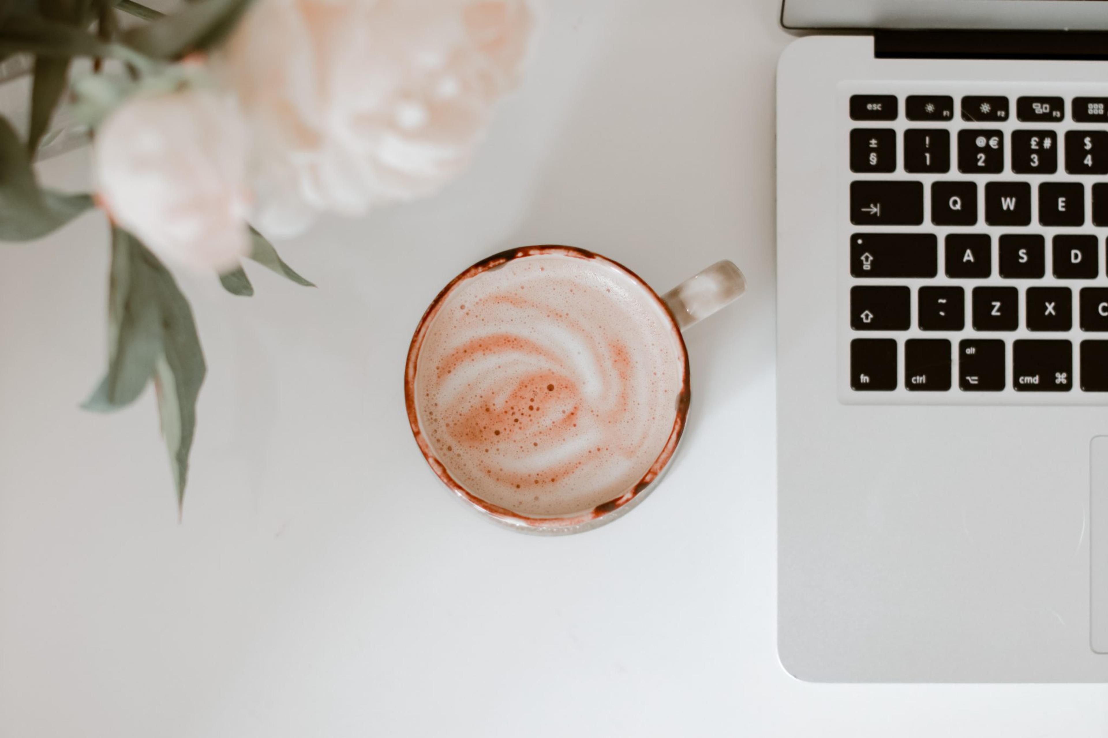 A frothy latte in a ceramic mug next to a laptop and blurred pink flowers on a white desk.