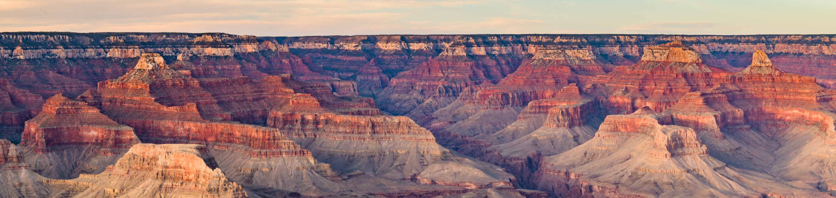 A vast expanse of layered red rock formations in closeup with a blurred background of the Grand Canyon under a soft, evening sky in Arizona.