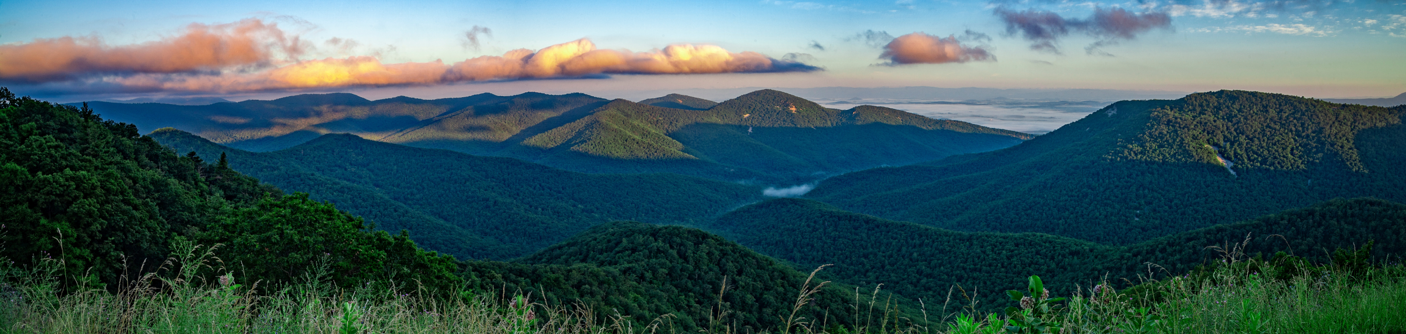 A panoramic view of the Blue Ridge Mountains in Virginia, with rolling green hills bathed in the warm light of a setting sun against a tranquil sky.