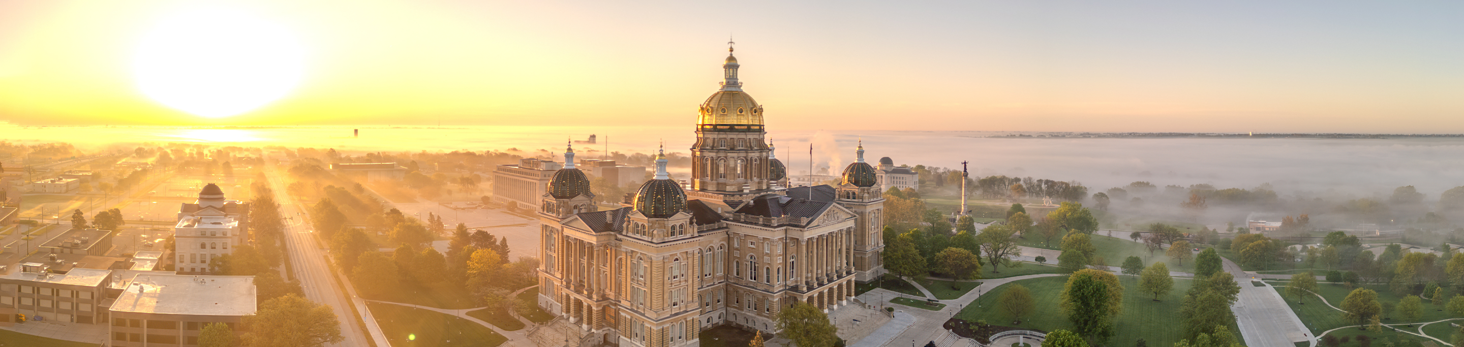 The Iowa State Capitol building in closeup with a blurred background of a misty morning sunrise casting golden light over Des Moines, Iowa.