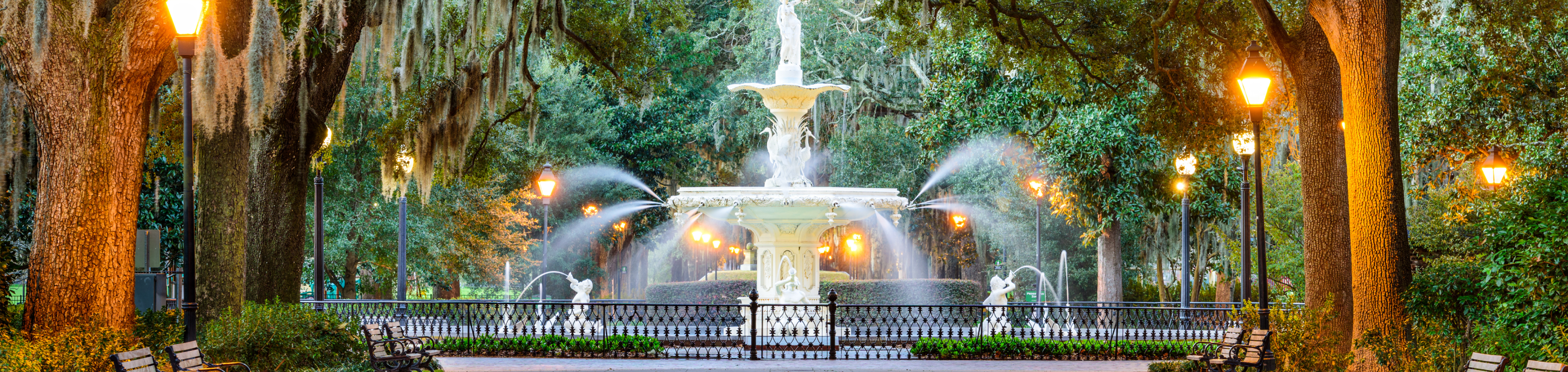 A historic fountain in closeup with a blurred background of oak trees draped in Spanish moss and glowing lampposts in Forsyth Park, Savannah, Georgia.