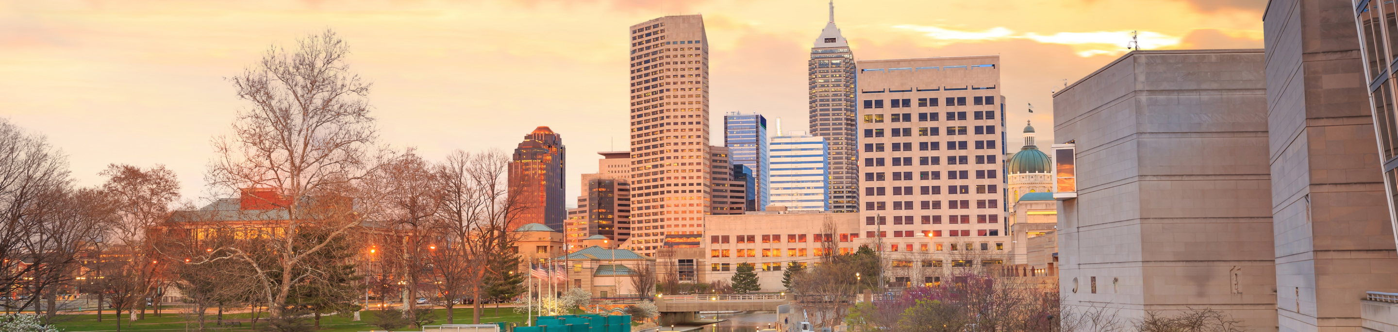 The Indianapolis skyline in closeup with a blurred background of leafless trees and the glowing lights of buildings under a warm sunset in Indiana.