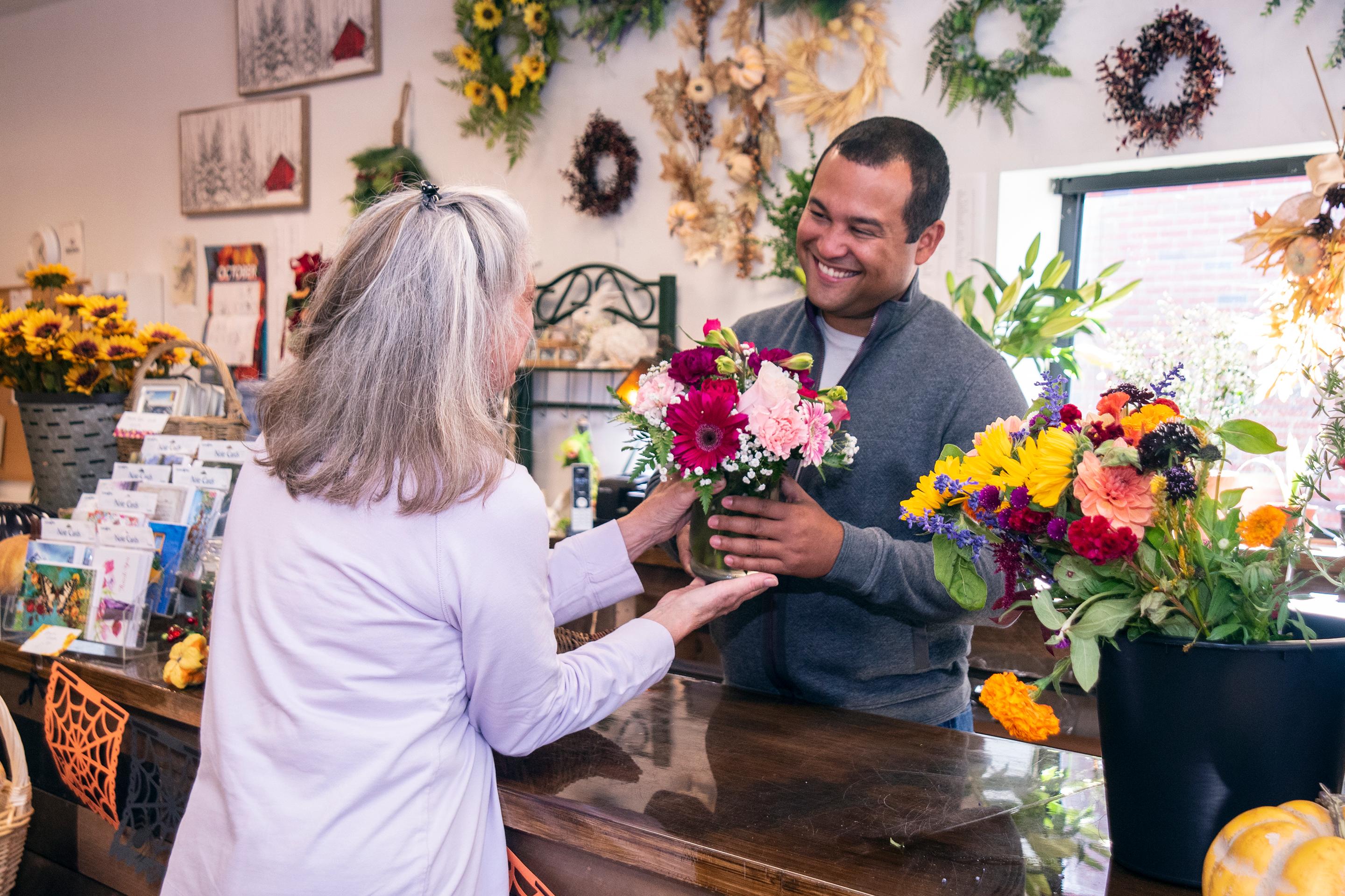 Image of Ken handing a bouquet to a woman purchasing flowers at flower shop counter