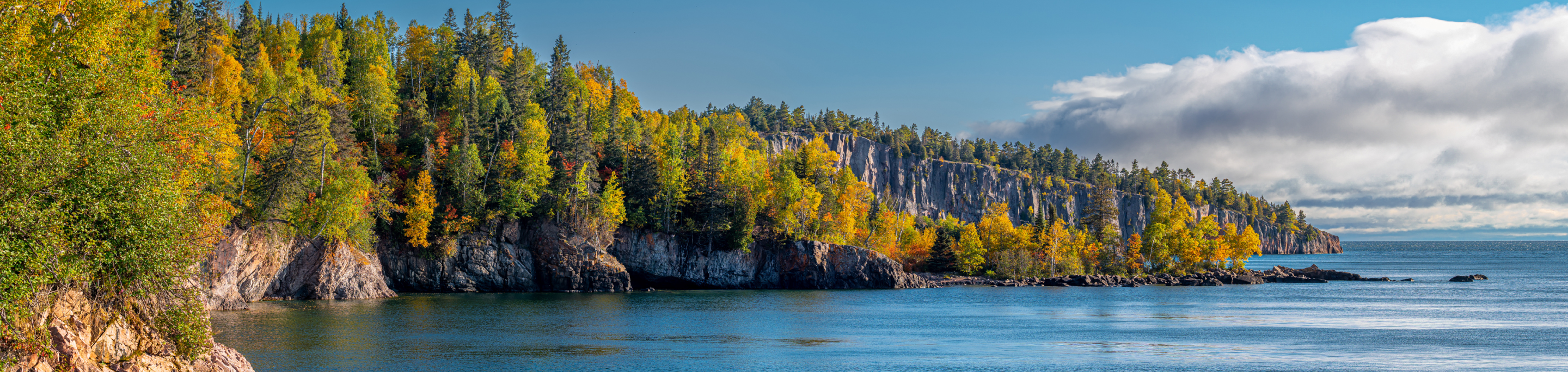 A rocky shoreline in closeup with a blurred background of vibrant autumn foliage and calm blue waters under a clear sky along the North Shore of Lake Superior in Minnesota.