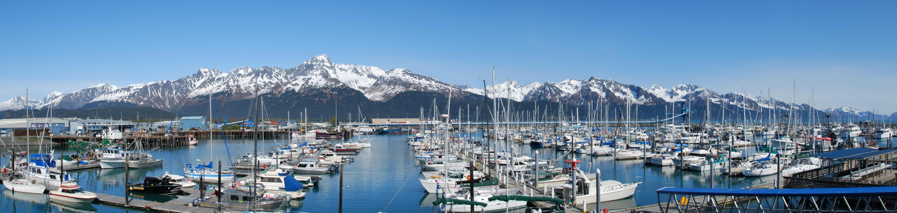 A marina filled with boats in closeup with a blurred background of snow-capped mountains under a clear blue sky in Seward, Alaska.