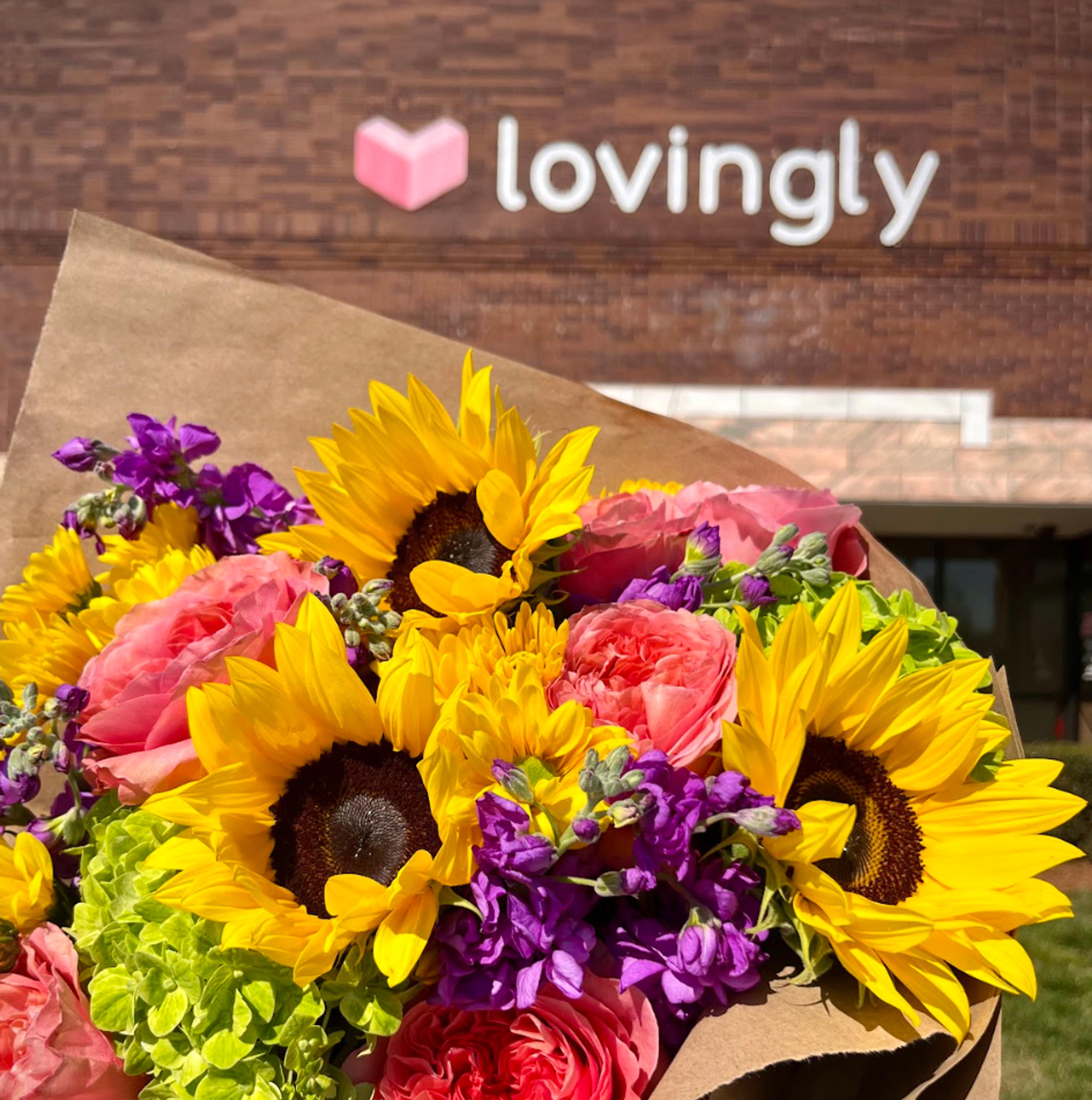 A colorful bouquet of sunflowers, roses, hydrangeas, and purple flowers, wrapped in brown paper, with the "lovingly" logo on a brick building in the background.
