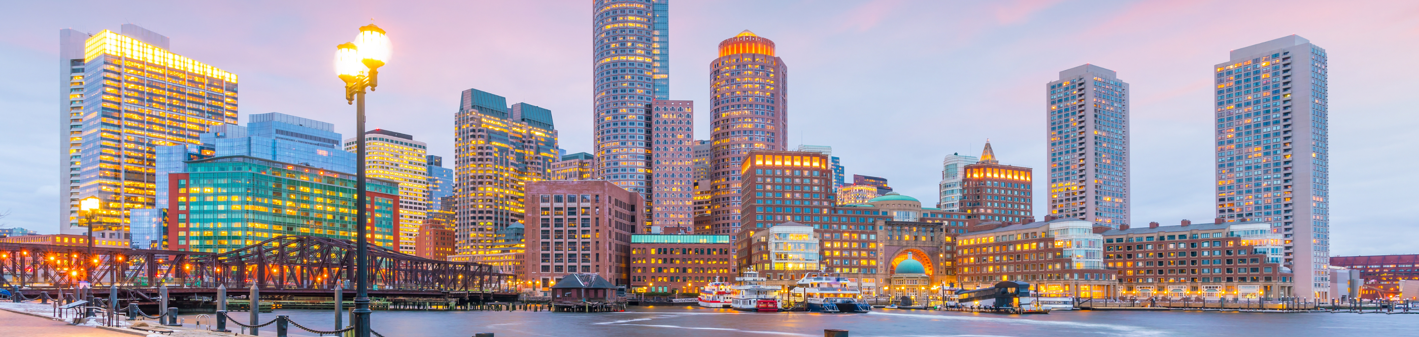 The Boston waterfront in closeup with a blurred background of illuminated skyscrapers and historic bridges under a twilight sky in Massachusetts.