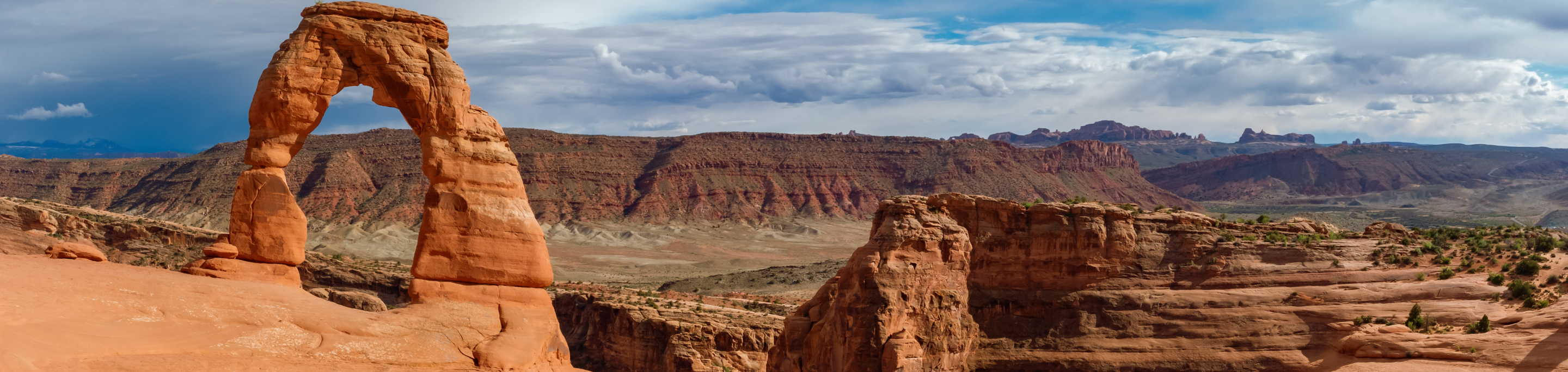 The iconic Delicate Arch stands tall against a backdrop of rugged red rock formations and distant mesas under a vast sky in Utah's Arches National Park.