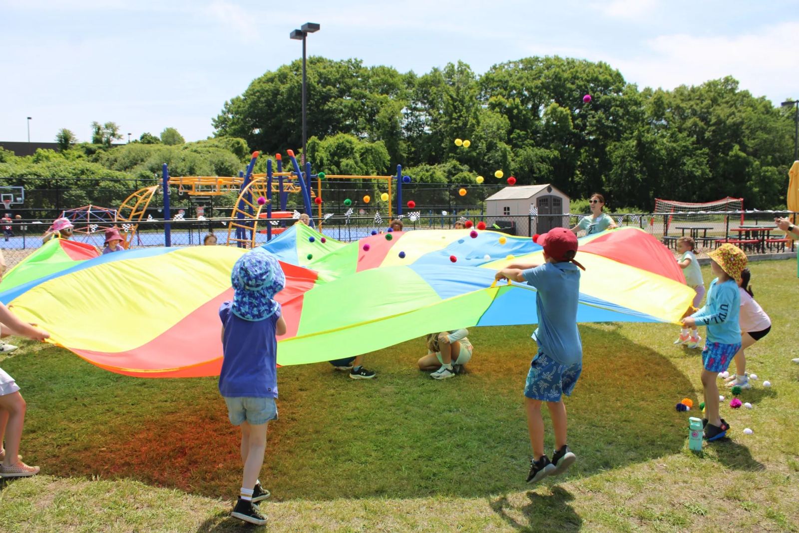 A group of SELA children play with a rainbow parachute outdoors on a sunny day.