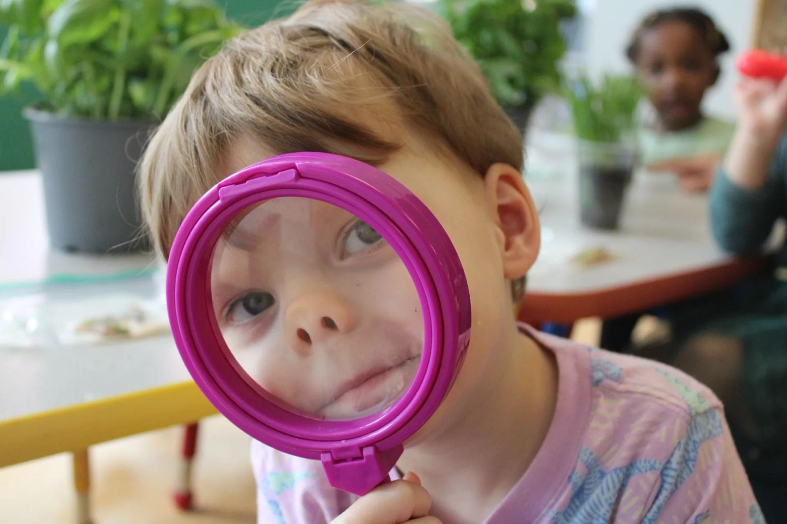 A SELA child holds a pink magnifying glass up to one eye, peering straight at the camera with a playful expression.