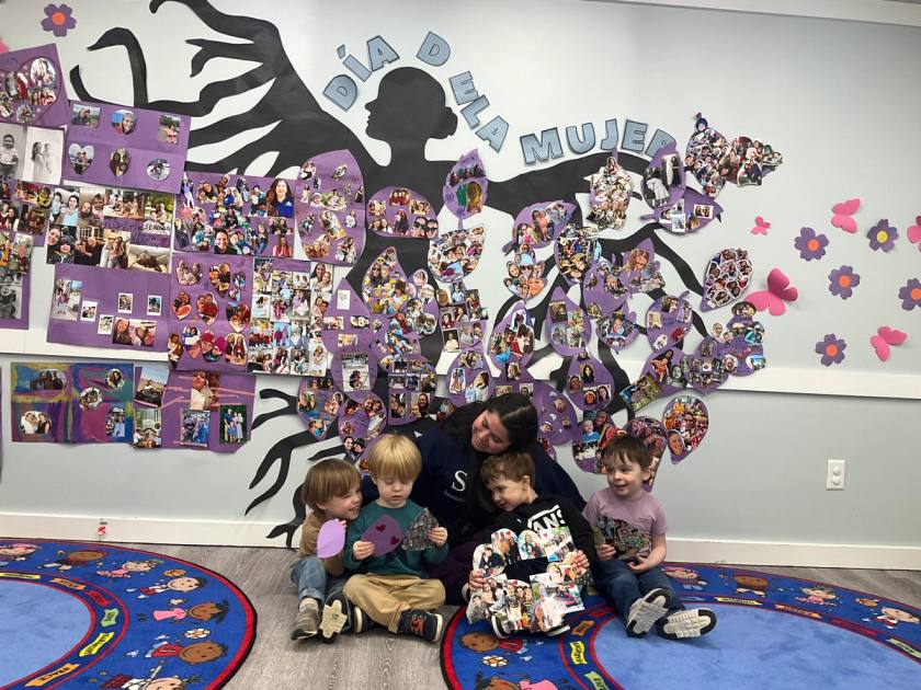 A teacher and four young students sitting together in front of a Día de la Mujer photo collage in a SELA classroom.
