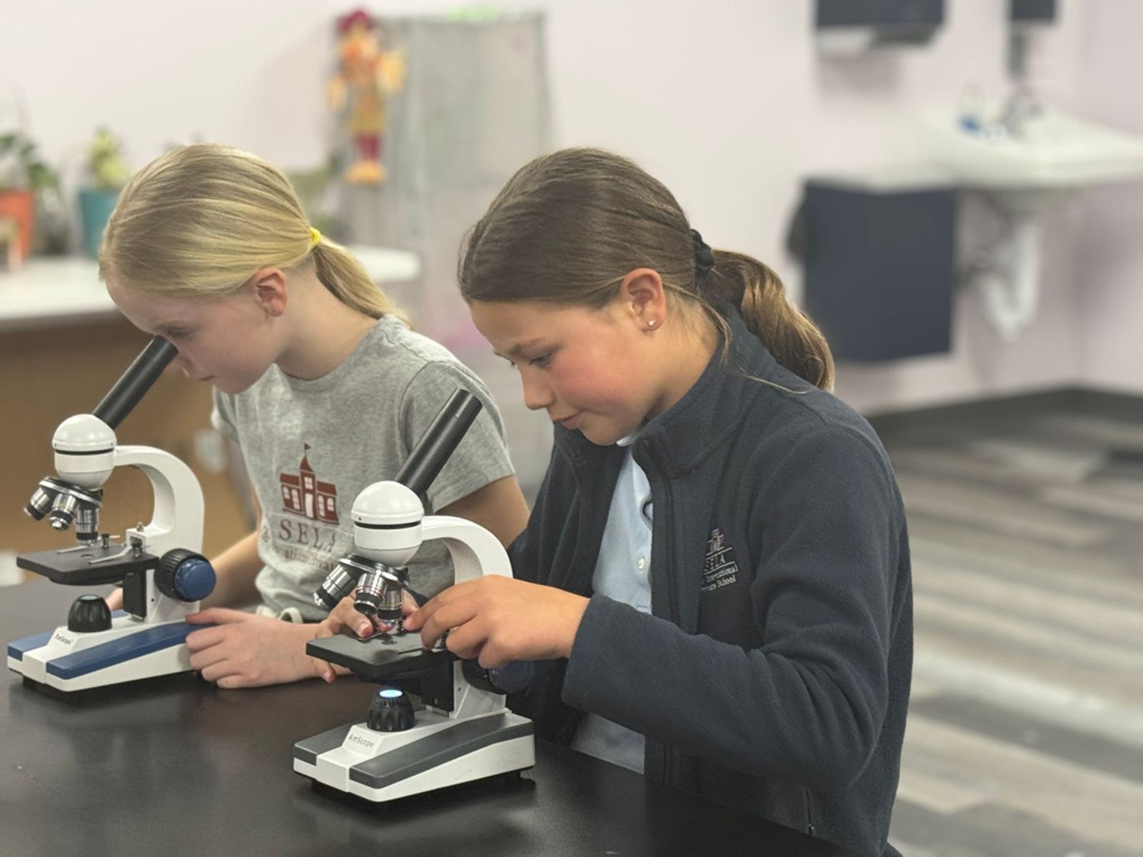 Two SELA Elementary students in the science lab looking at bacteria under a microscope