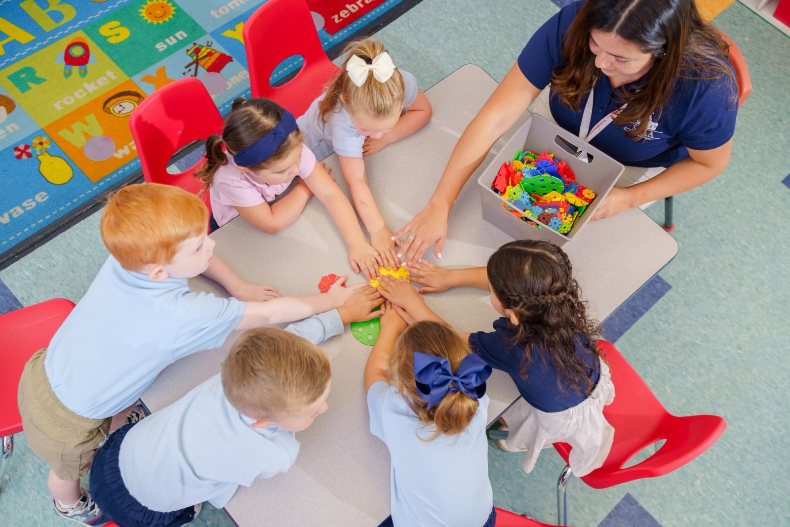 A circle of SELA children stand together in their bright classroom holding stuffed animals and toys, mid-laugh during morning meeting.