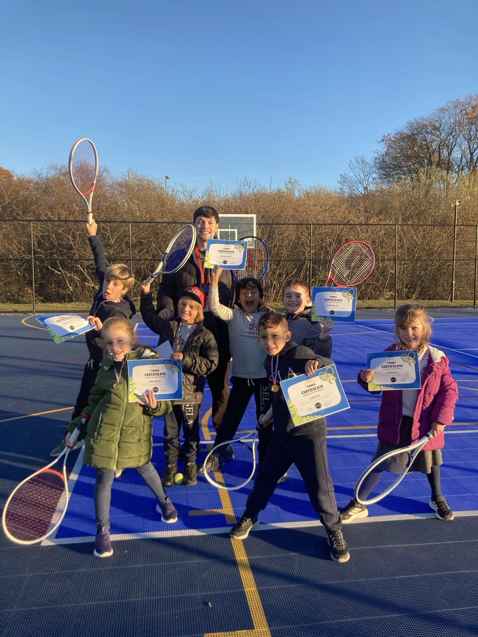 A group of SELA children hold up tennis certificates on the blue court in warm late-afternoon sunlight.