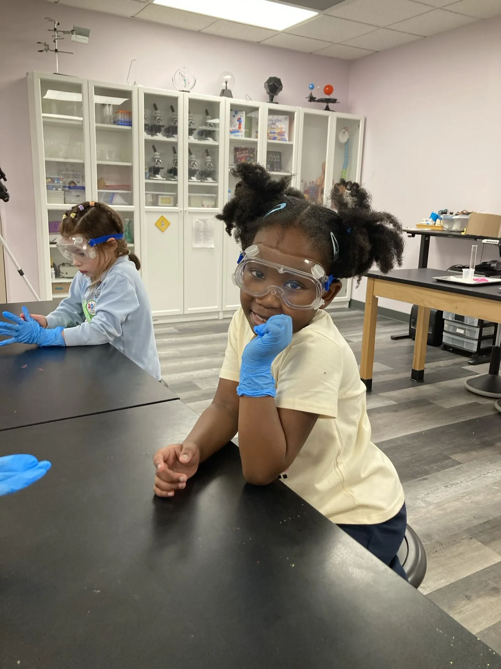 A SELA student in safety goggles and blue gloves grins at a lab table during a science enrichment session.
