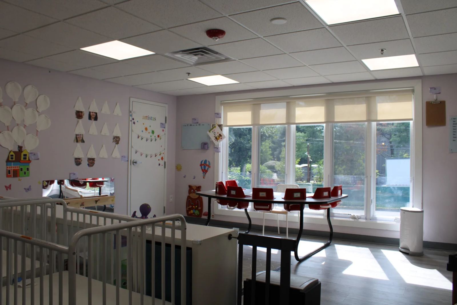 An early-years classroom at SELA's Norwell campus, with cribs and a sunlit activity table by large windows.