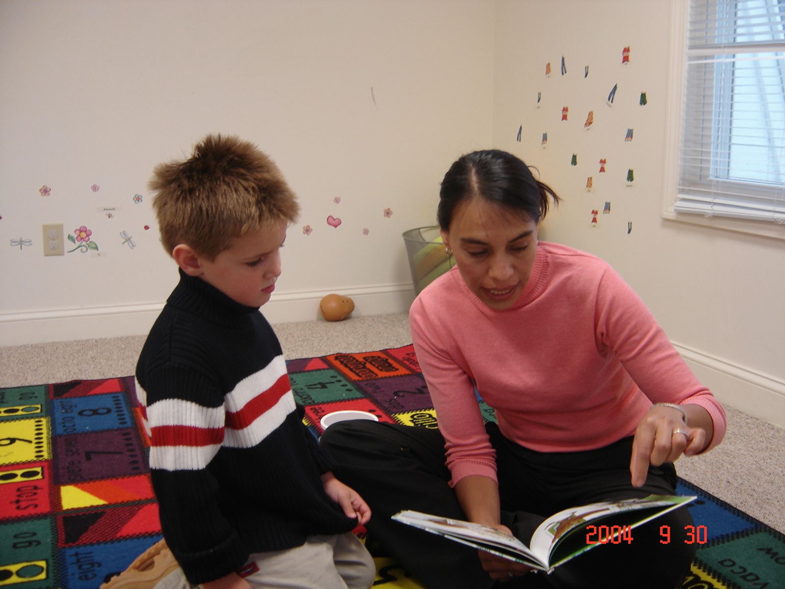 Young SELA students gather around a classroom table for a group painting activity, with Spanish day-of-the-week cards displayed on the wall behind them.
